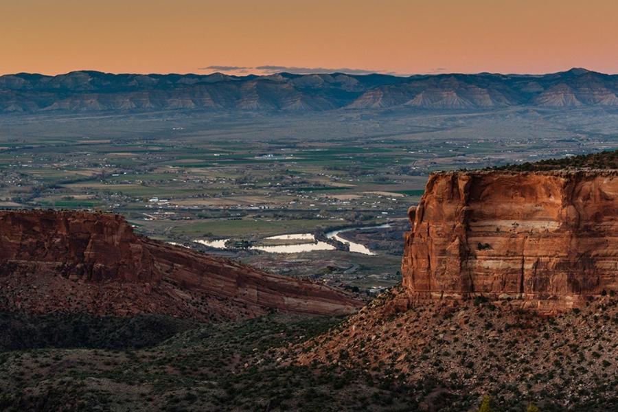 An aerial view of a large outcropping of reddish rock in Colorado National Monument, overlooking the Town of Fruita and distant mountains.