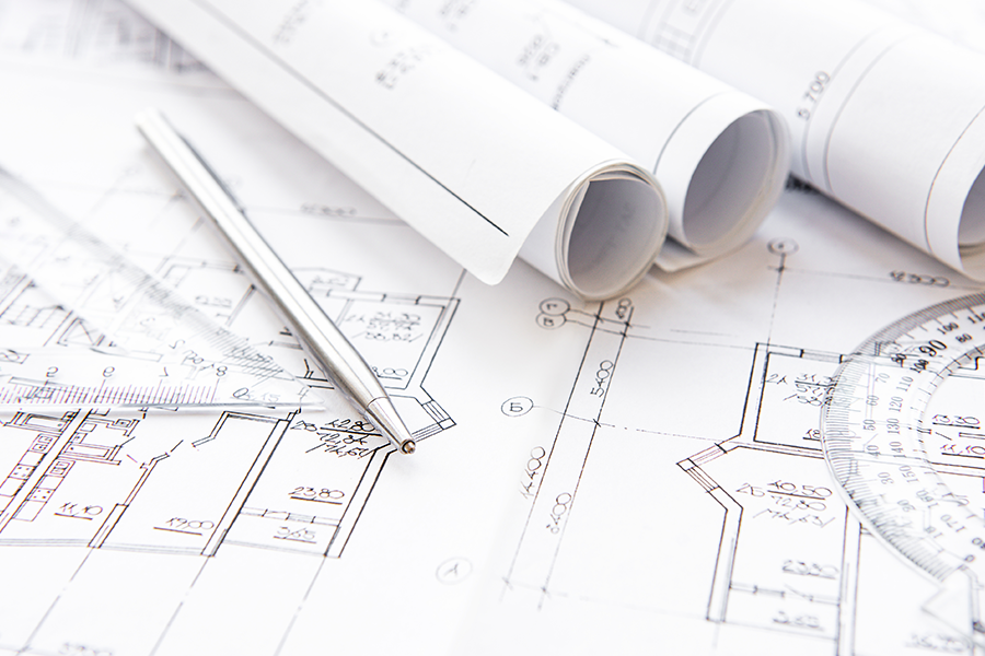 Rolled architectural blueprints lie on top of detailed building plans, with a metal pen and measuring tools placed on the drawings.